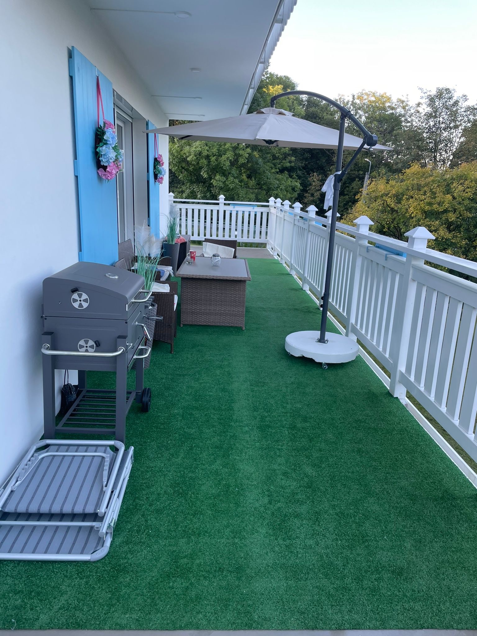 Zadaszony balkon z sztuczną trawą, białymi balustradami, szarym grillem, rozłożonym leżakiem, parasolem i rattanowymi meblami wypoczynkowymi, ozdobiony niebieskimi okiennicami z kwiatowymi...