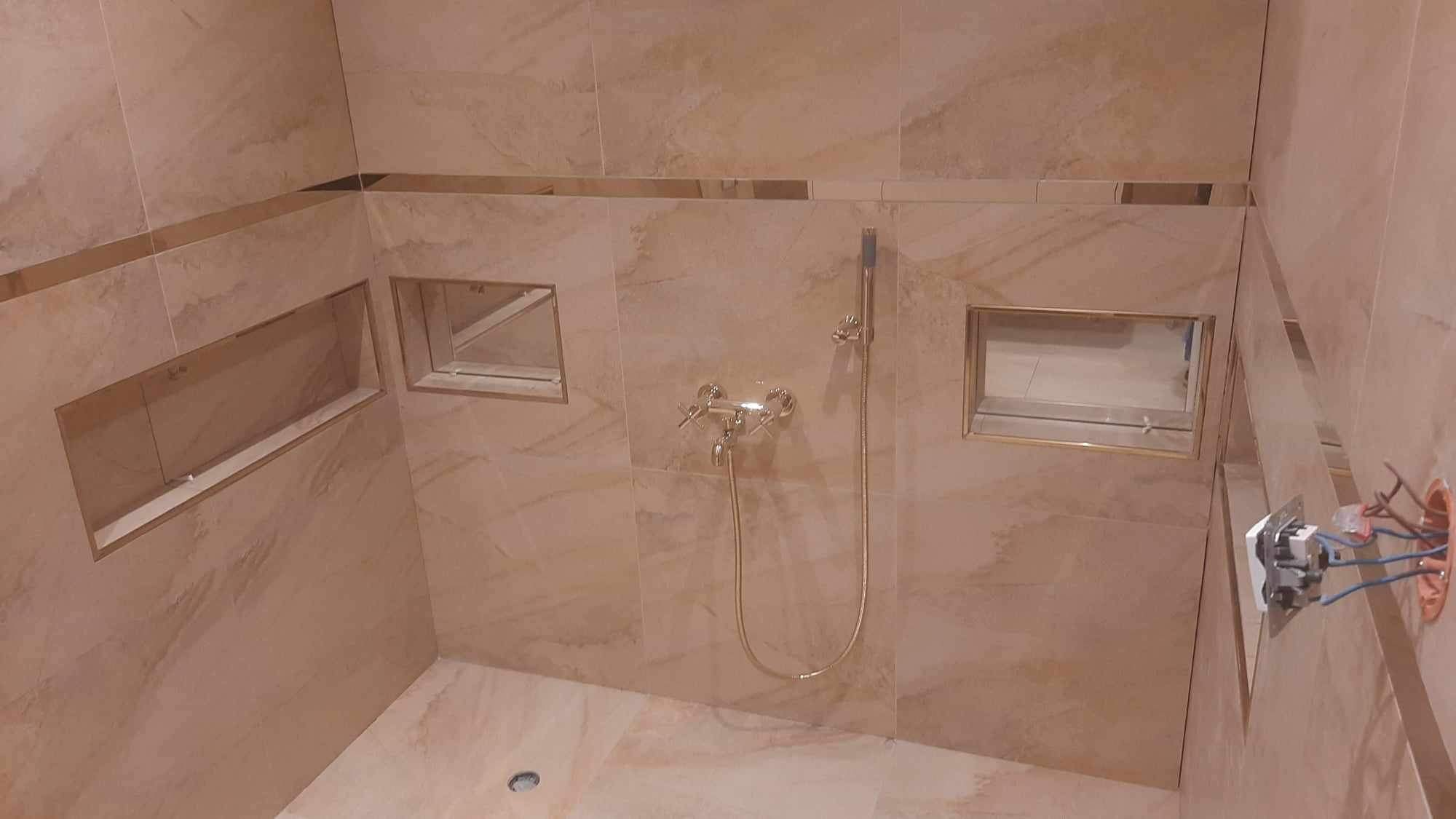 Wykończona kabina prysznicowa z beżowych płytek imitujących marmur w Jeleniej Górze, z wnękami na kosmetyki wykończonymi złotą listwą. Widoczna bateria prysznicowa i wystająca instalacja elektryczna.
