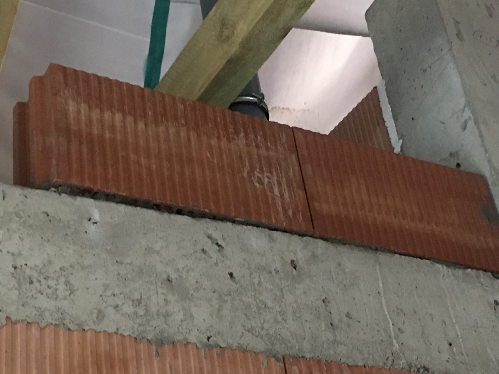 Fragment ściany z czerwonej cegły, z widoczną warstwą betonu i fragmentem rury oraz drewnianą belką, ukazujący etap prac budowlanych.