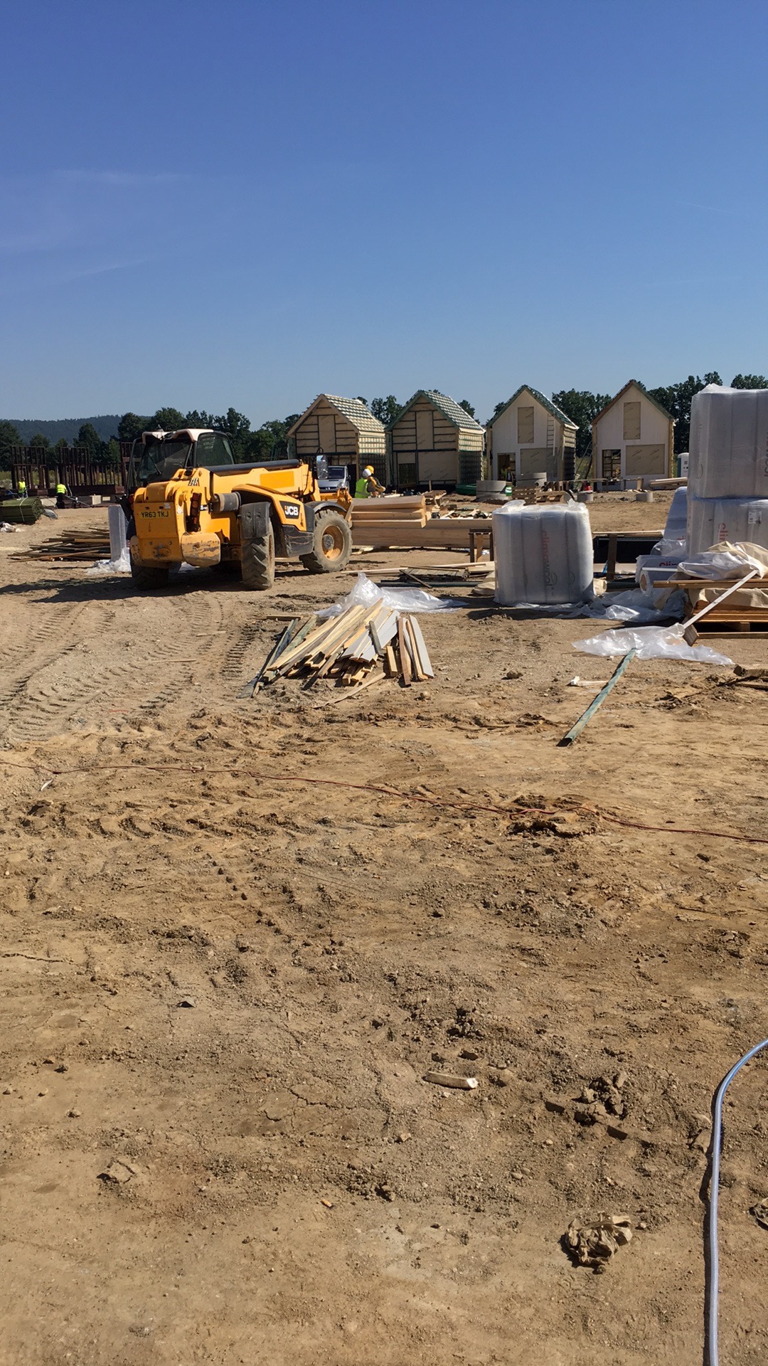 Budowa domów jednorodzinnych w stanie surowym otwartym, widoczny żółty wózek widłowy JCB, materiały budowlane i pracownicy w oddali.