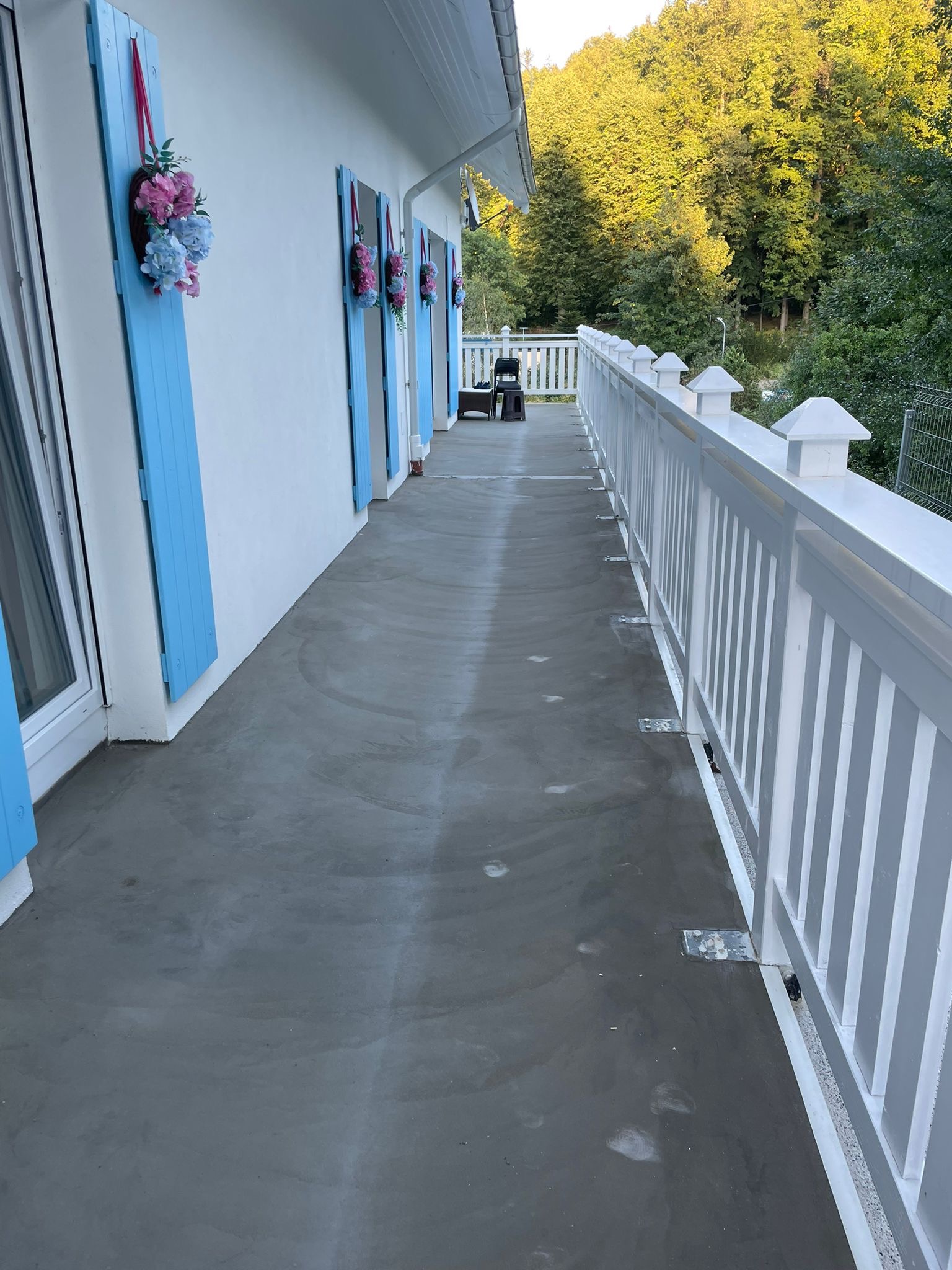 Długi balkon z nową, gładką wylewką betonową, białą balustradą i błękitnymi okiennicami ozdobionymi kwiatowymi dekoracjami.