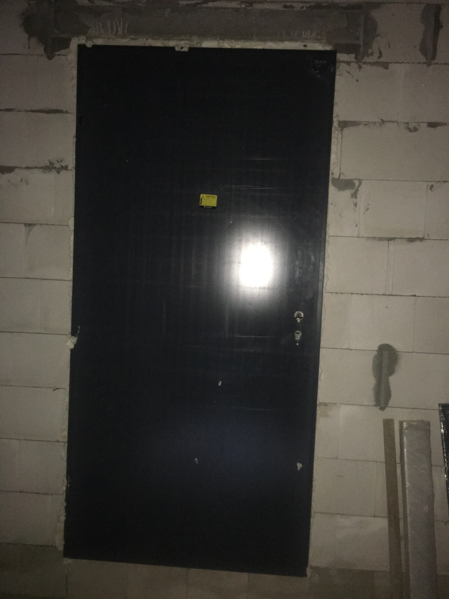 Czarna, częściowo zamontowana, antywłamaniowa  drzwi wejściowe w surowym otworze ściany z szarych bloczków betonowych; widoczne ślady zaprawy montażowej.