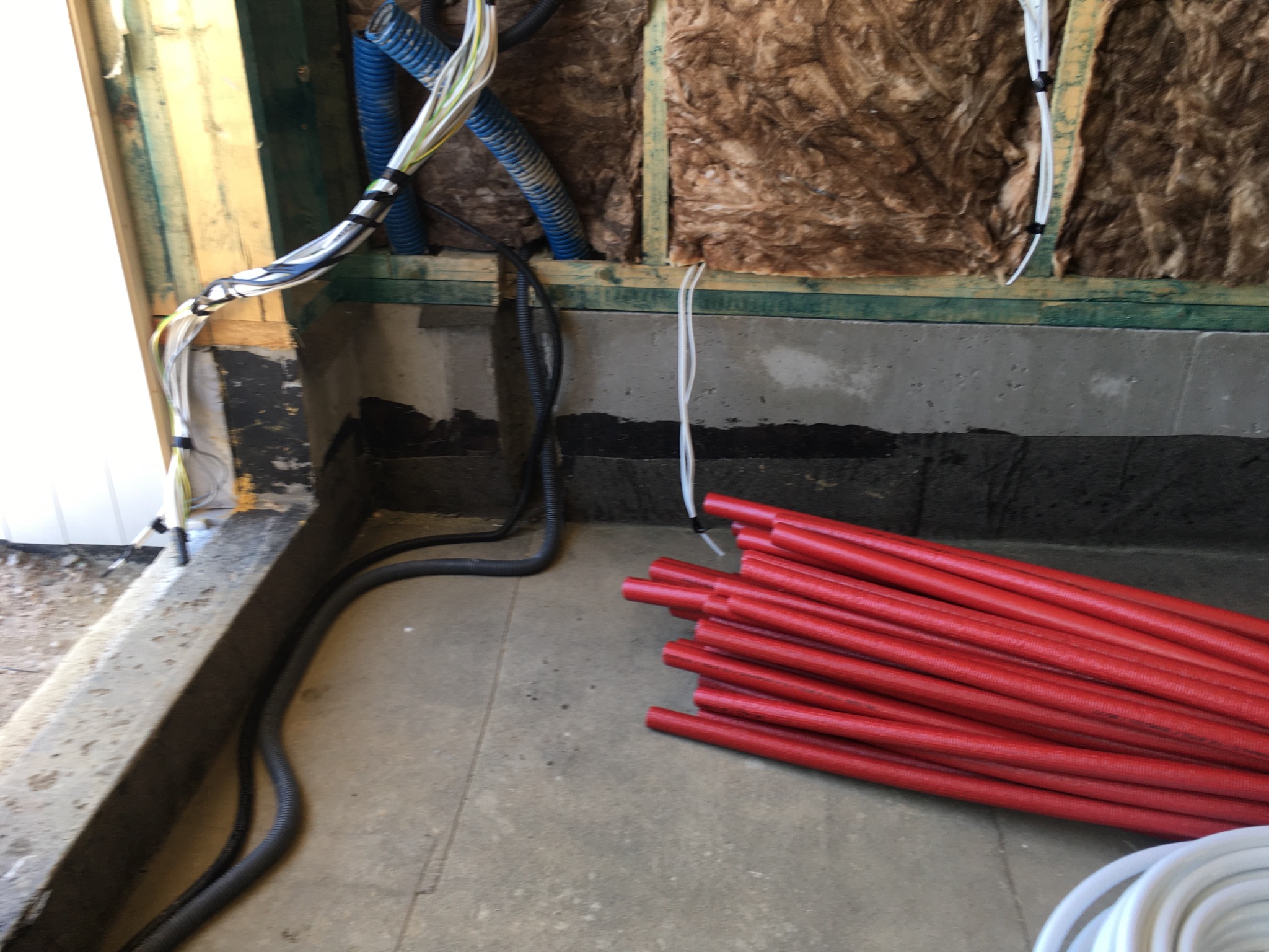 Surowy stan ścian z izolacją z wełny mineralnej, widoczne przewody elektryczne, rury w kolorze czerwonym i niebieskim oraz czarne peszle na podłodze w trakcie prac wykończeniowych.