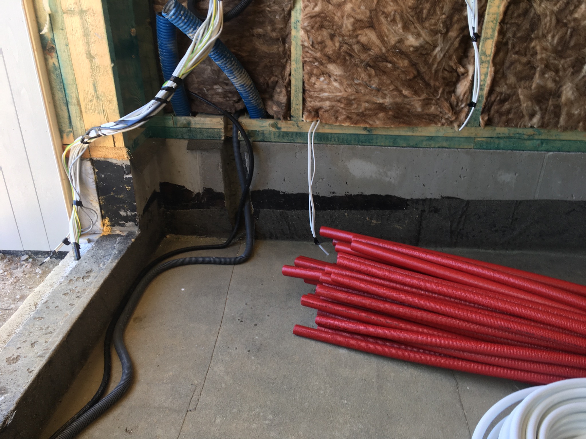 Ściana w trakcie prac instalacyjnych: widoczne przewody elektryczne w peszlach, izolacja z wełny mineralnej, oraz stos czerwonych rur karbowanych na podłodze.