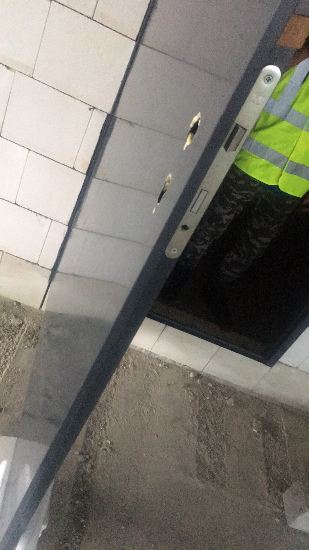 Montaż ościeżnicy drzwiowej z użyciem poziomicy; w tle widoczna osoba w kamizelce odblaskowej i fragment podłogi w stanie surowym.