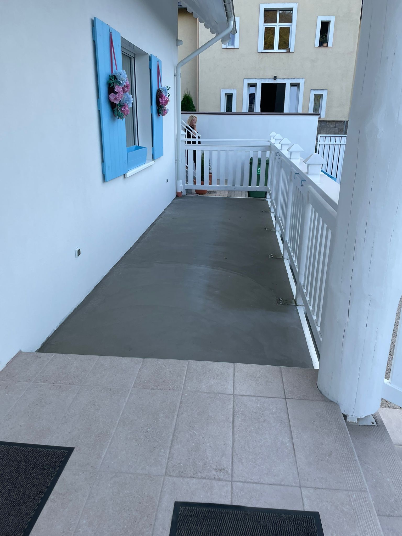Wylewka betonowa na balkonie z białymi balustradami i niebieskimi okiennicami z dekoracjami kwiatowymi, widok z poziomu wejścia na balkon.