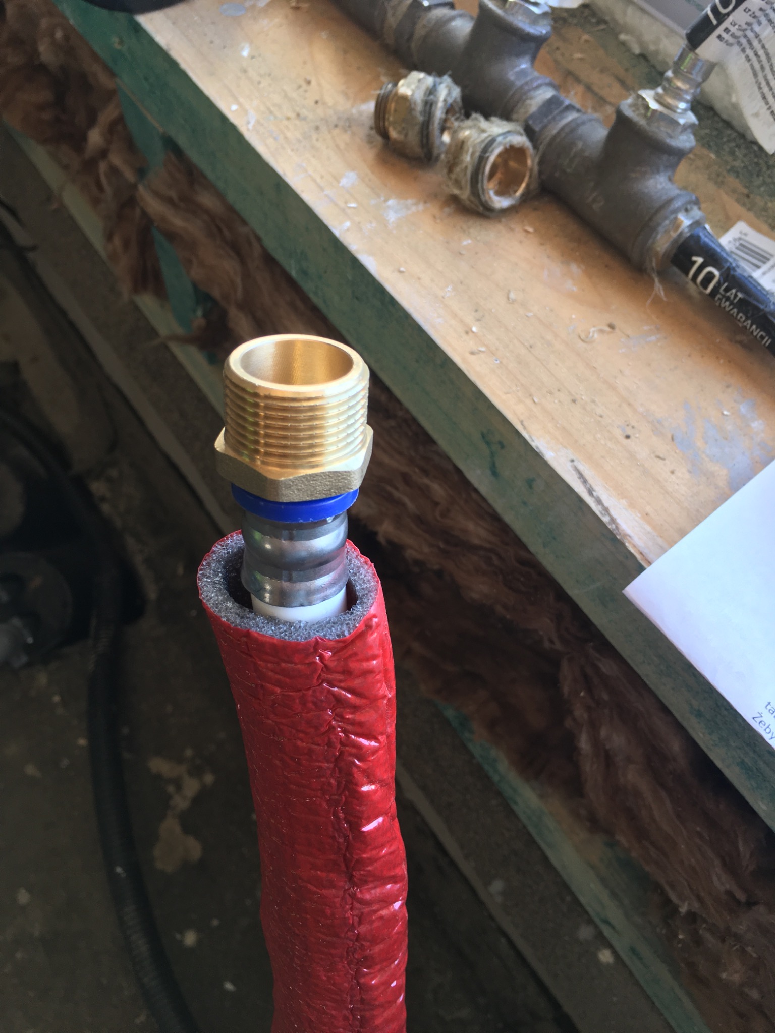 Fragment instalacji centralnego ogrzewania: rura w czerwonej izolacji z mosiężnym gwintem i niebieską uszczelką, na tle drewnianej konstrukcji z widoczną wełną mineralną.