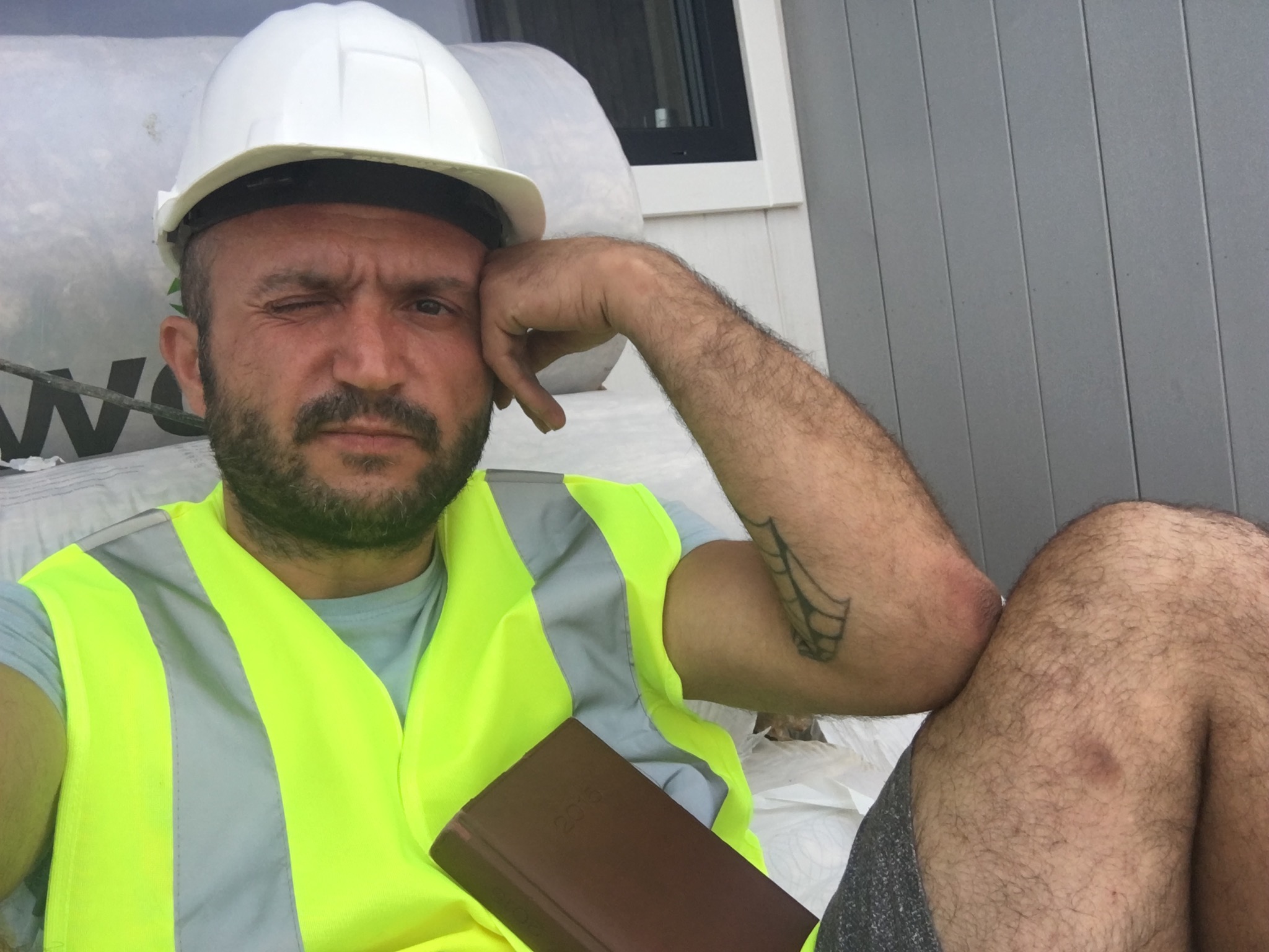 Pracownik budowlany w kasku i kamizelce odblaskowej, z przymrużonym okiem, oparty o rękę, z kalendarzem w dłoni, na tle folii i szarej ściany.