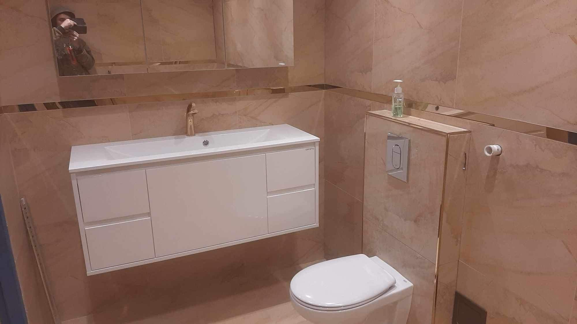 Nowoczesna łazienka z beżowymi płytkami imitującymi marmur, białą podwieszaną szafką z umywalką i złotą armaturą, toaleta podwieszana, odbicie fotografa w lustrze.