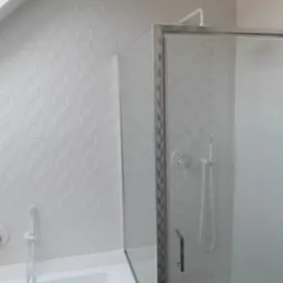 Nowoczesna, jasna łazienka w odcieniach bieli z kabiną prysznicową ze szklanymi drzwiami i deszczownicą, oraz wanną z baterią i słuchawką, wykończona płytkami z subtelnym wzorem jodełki na ścianach.