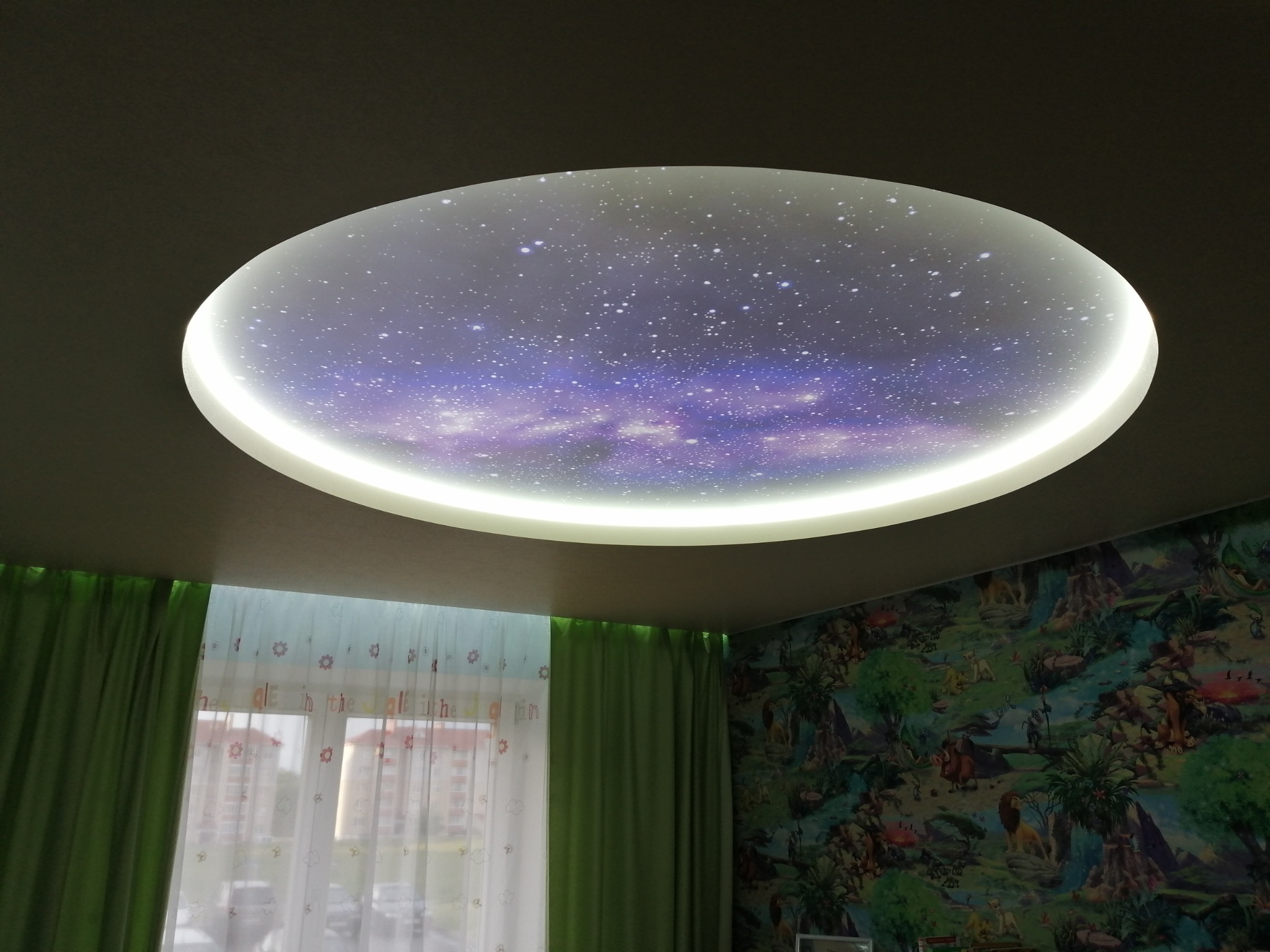 Okrągły sufit napinany z motywem kosmosu, oświetlony od wewnątrz, w pokoju z zasłonami i tapetą w dżunglę. Oryginalny design sufitu w pokoju dziecięcym.