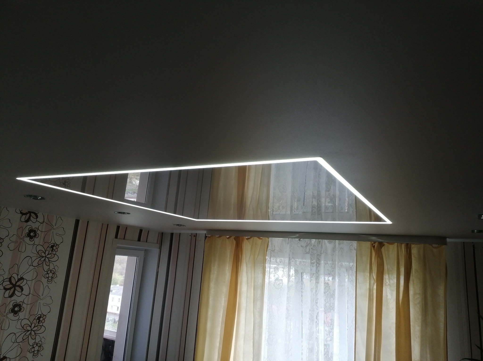 Nowoczesny sufit z połyskiem i oświetleniem LED w kształcie trapezu, odbijający okno i firanki. Widok z dołu na wykończone wnętrze.