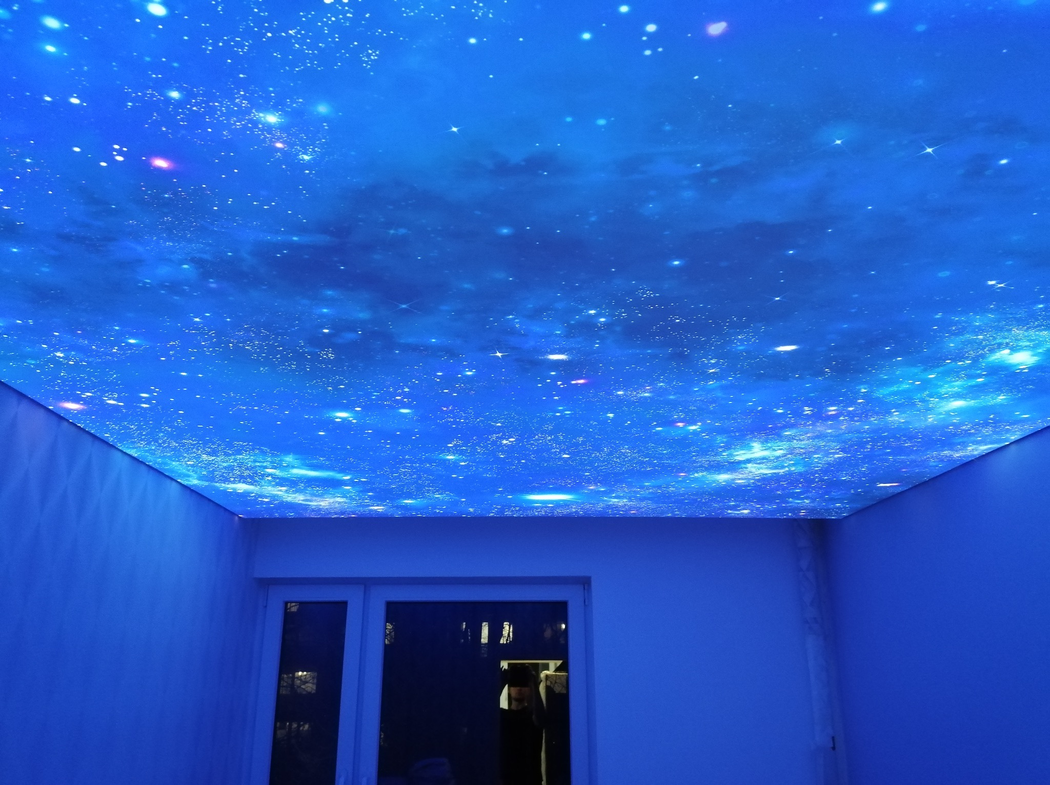 Pokój z efektem gwieździstego nieba na suficie. Błękitne oświetlenie podkreśla kosmiczny wzór. Widoczne drzwi i białe ściany.