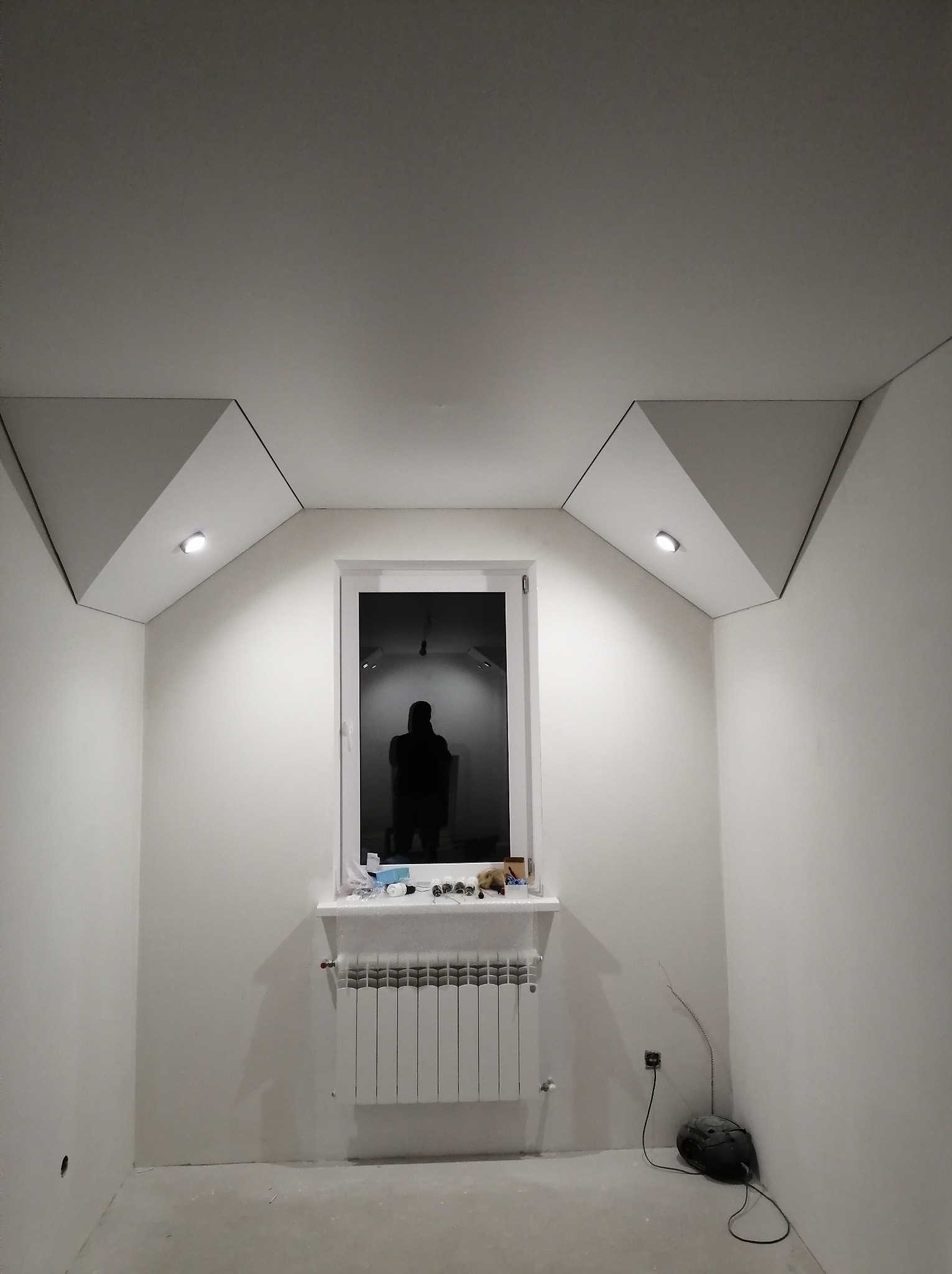 Białe wnętrze z oknem, grzejnikiem i nowym sufitem z podwieszanymi elementami i wbudowanymi lampkami. Widoczny cień osoby w oknie.