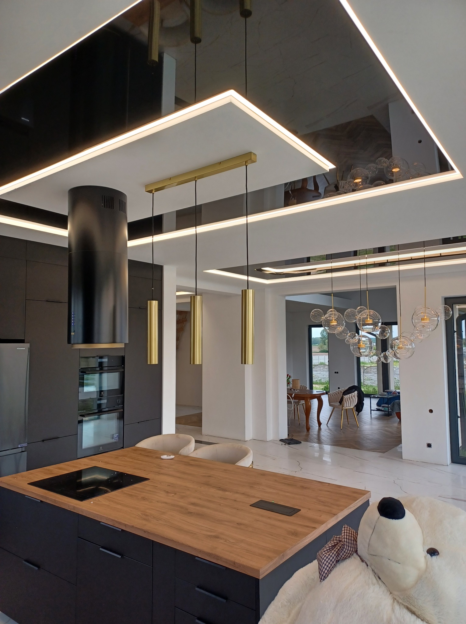 Nowoczesny salon z czarnym sufitem napinanym z oświetleniem LED, odbijającym detale wnętrza, w tym wyspę kuchenną i lampy wiszące.