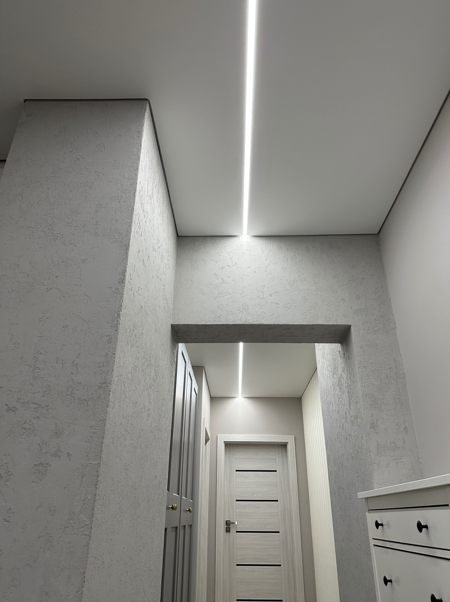 Nowoczesny korytarz z oświetleniem liniowym LED w suficie i dekoracyjną strukturą ścian. Widoczne drzwi i komoda w stonowanej kolorystyce.