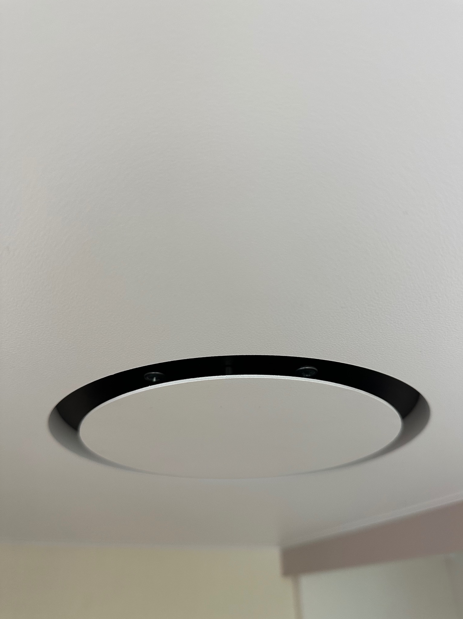 Biały sufit z okrągłym, wpuszczonym panelem oświetleniowym z czarną obwódką. Minimalistyczny design, nowoczesne oświetlenie w pomieszczeniu.