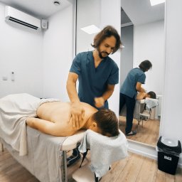 Vital Massages For Health - Mężczyzna w niebieskim uniformie masuje plecy pacjenta leżącego na stole do masażu w jasnym, minimalistycznym gabinecie. Widoczne odbicie w lustrze ukazuje dalszą część pomieszczenia.