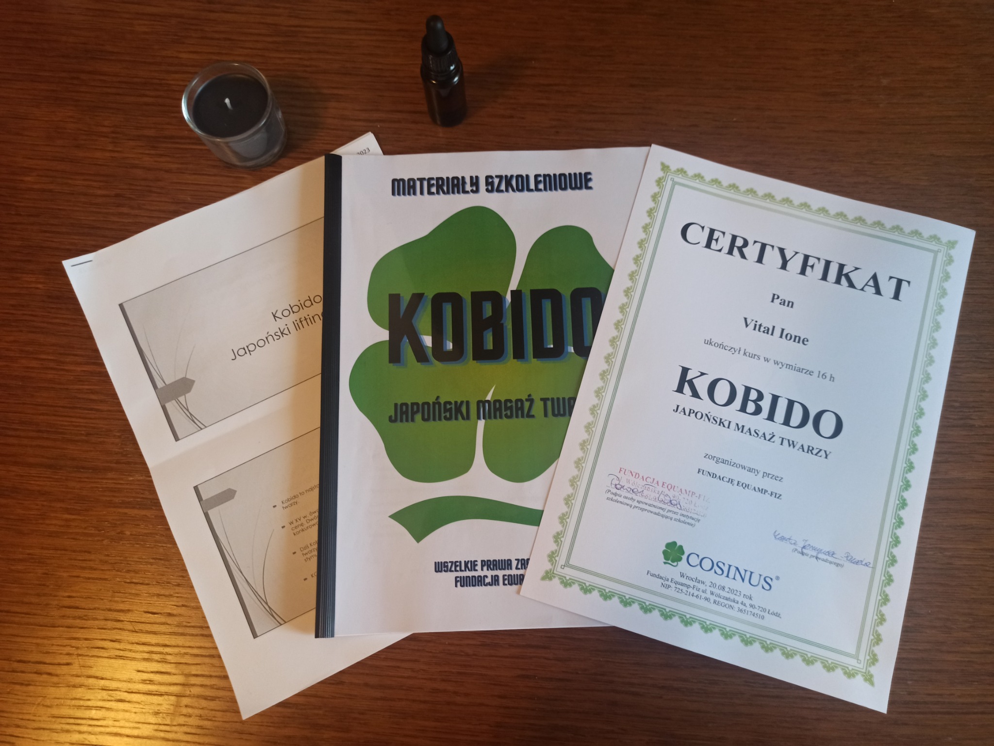 Materiały szkoleniowe, certyfikat ukończenia kursu i olejek do masażu Kobido, japońskiego masażu twarzy, na drewnianym stole, obok świeczka.