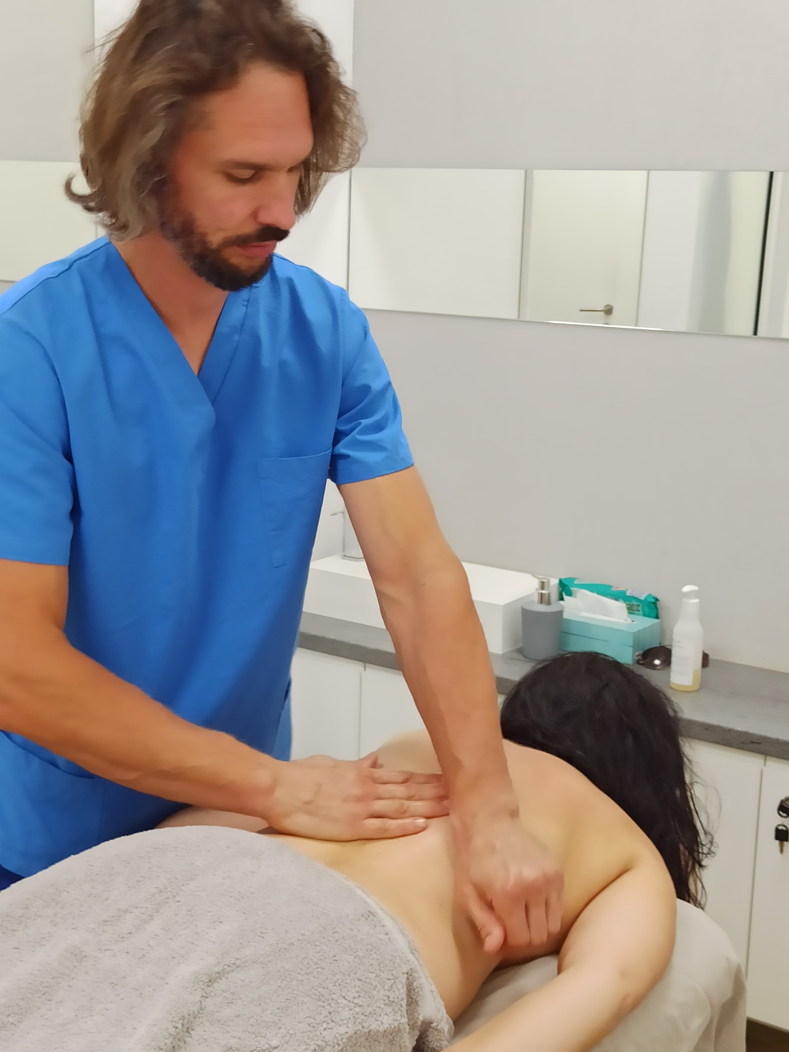 Mężczyzna w niebieskim uniformie wykonuje masaż pleców pacjentki leżącej na leżance przykrytej szarym ręcznikiem, w tle białe szafki i lustro.