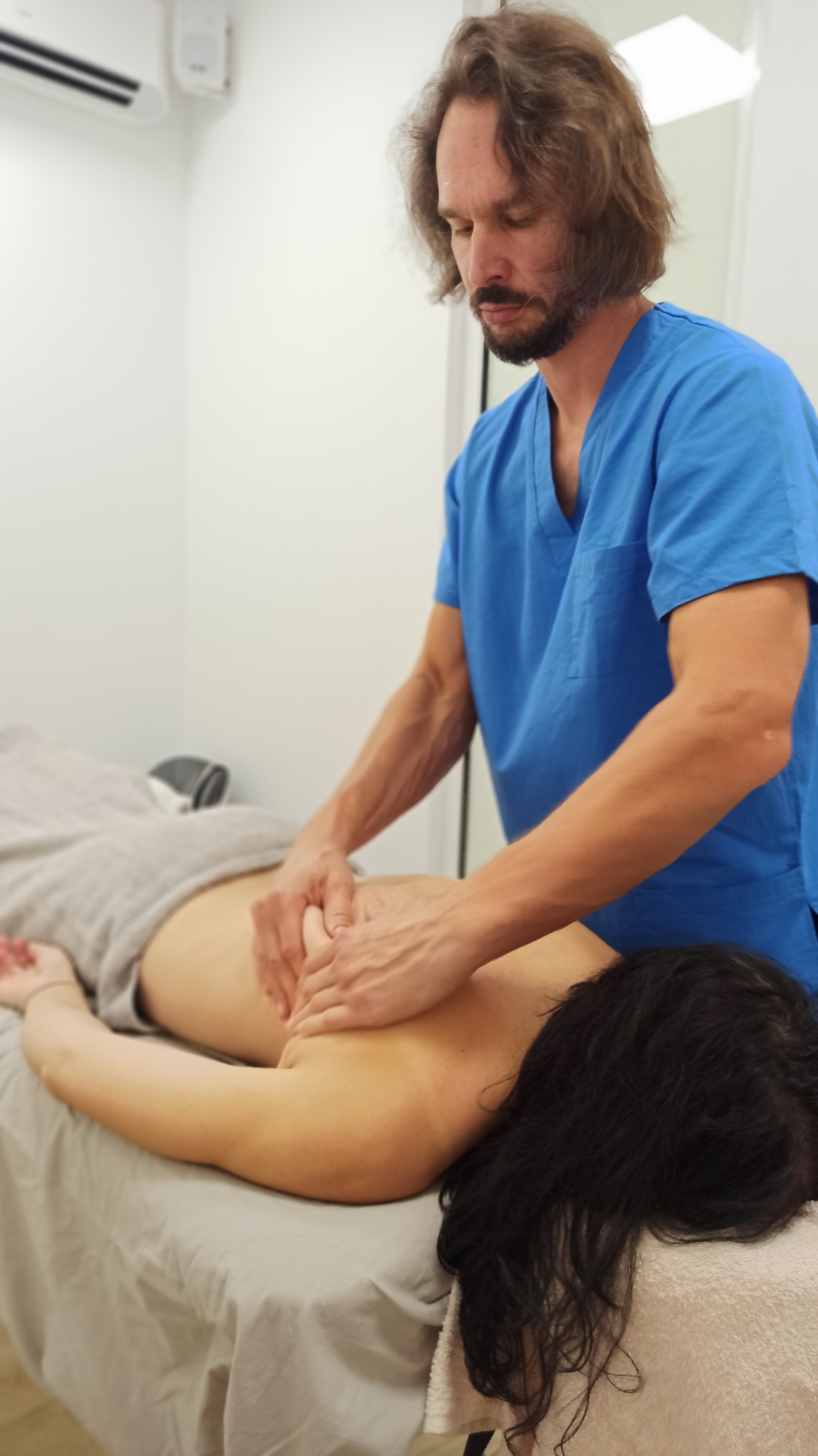 Mężczyzna w niebieskim uniformie wykonuje masaż pleców pacjentki leżącej na stole w jasnym gabinecie.