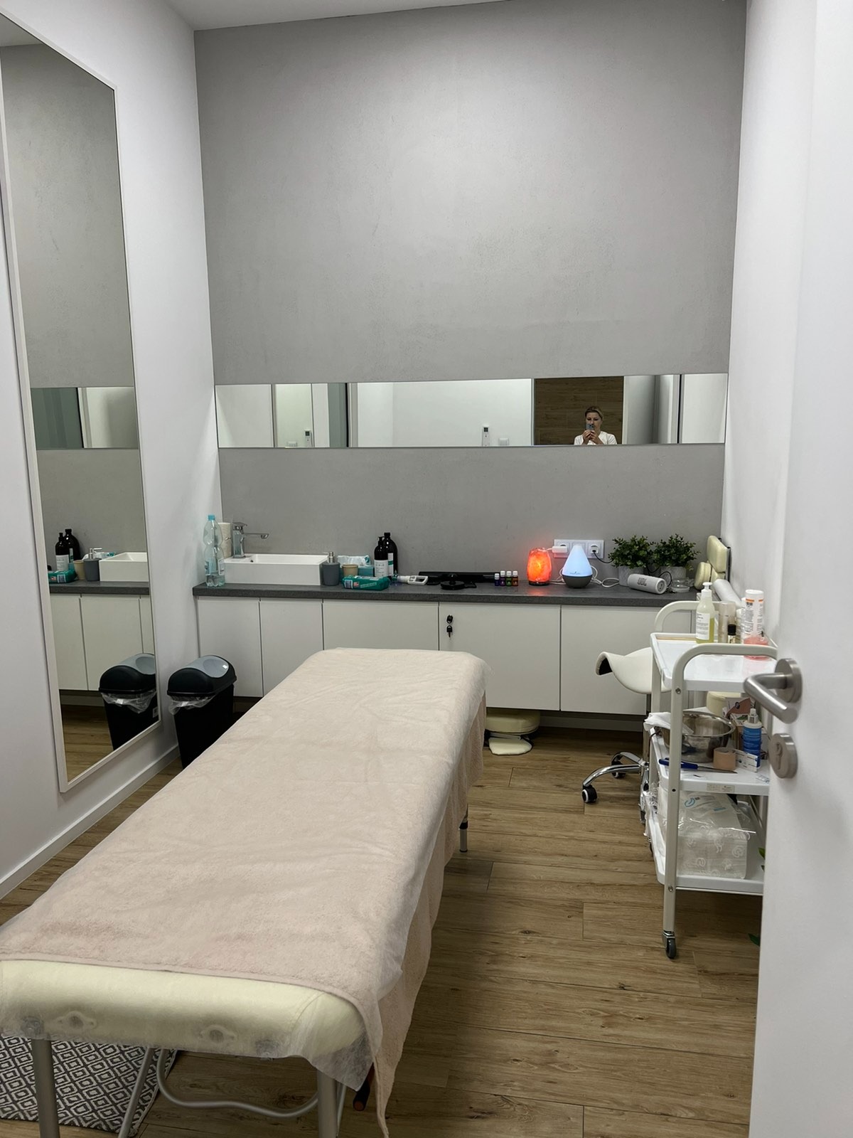 Jasne wnętrze gabinetu z łóżkiem do zabiegów, szafką z umywalką, lustrami i wózkiem z akcesoriami, widoczny dyfuzor zapachowy.