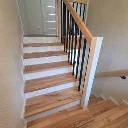 Stolarstwo Andrzej Robiński - Jasne, drewniane schody z białymi podstopnicami i czarną balustradą, widziane z góry. Minimalistyczny design, naturalne światło, tekstura ściany w tle.