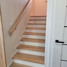 Stolarstwo Andrzej Robiński - Jasne, drewniane schody z białymi podstopnicami i drewnianą poręczą w domu w Górze. Nowoczesny design, minimalistyczne wykończenie, widok z dołu.