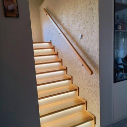 Stolarstwo Andrzej Robiński - Jasne, drewniane schody z podświetleniem LED, zamontowane przy ścianie o fakturze baranka. Drewniana poręcz. Widok z dołu, z boku. Nowoczesny design.