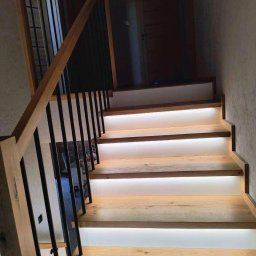 Stolarstwo Andrzej Robiński - Nowoczesne schody z drewna z podświetleniem LED, czarne metalowe barierki. Jasne drewno, minimalistyczny design, ciepłe światło. Detal wykończenia wnętrza.