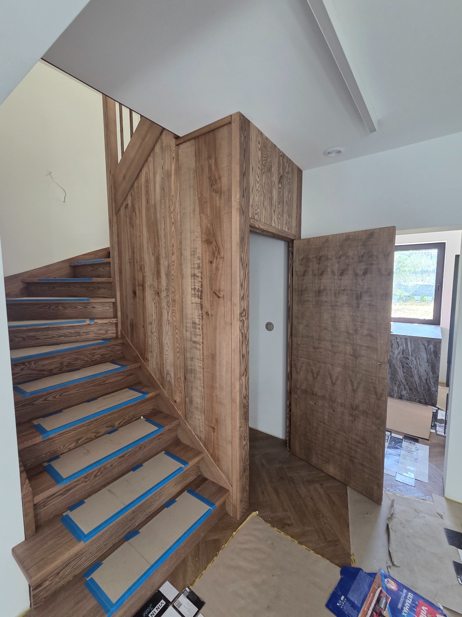 Wykończone drewniane schody z zabezpieczonymi stopniami, dopasowane kolorystycznie do drewnianej boazerii i drzwi w nowoczesnym wnętrzu domu.