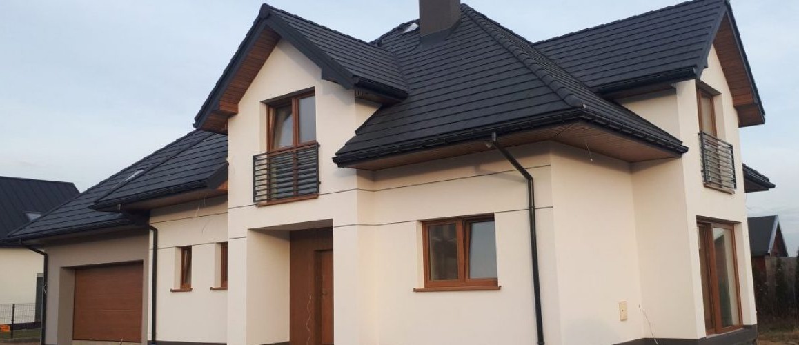 Dwukondygnacyjny dom z ciemną dachówką, jasną elewacją i drewnianymi oknami oraz garażem po lewej stronie.