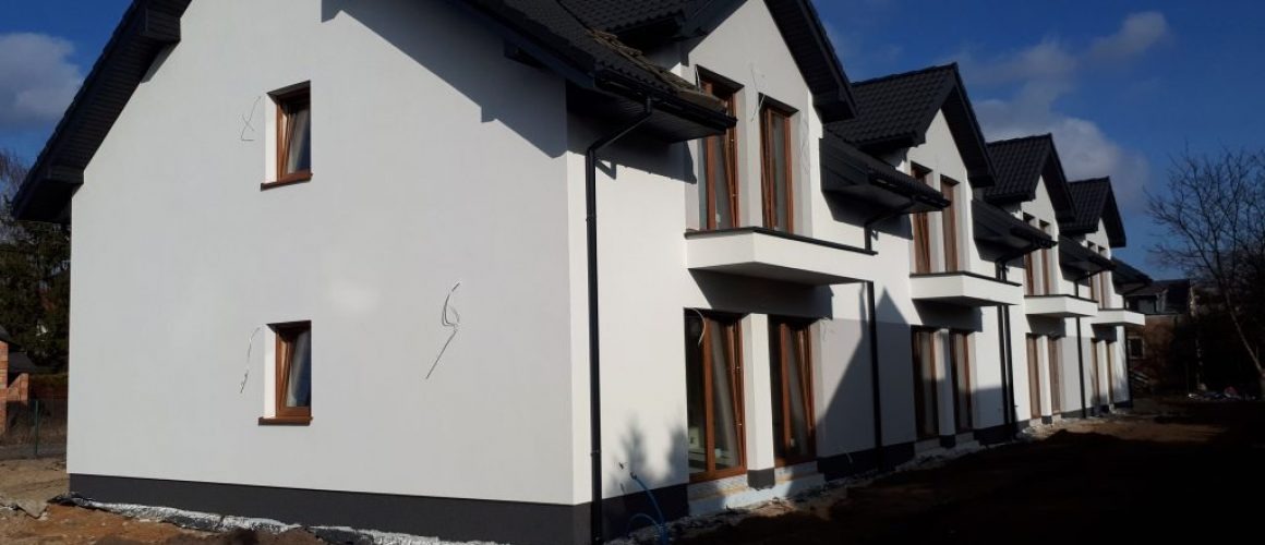 Szereg identycznych, nowo wybudowanych domów z białymi elewacjami, brązowymi oknami i ciemnymi dachami, widoczne wystające kable na ścianie jednego z domów.
