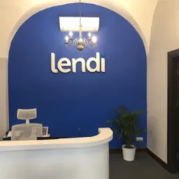 Recepcja biura z białym biurkiem, krzesłem z siatkowym oparciem, rośliną w doniczce i logo 'lendi' na niebieskiej ścianie pod łukowatym sufitem.