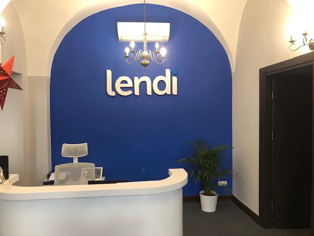 Recepcja biura z białym biurkiem, krzesłem z siatkowym oparciem, rośliną w doniczce i logo 'lendi' na niebieskiej ścianie pod łukowatym sufitem.