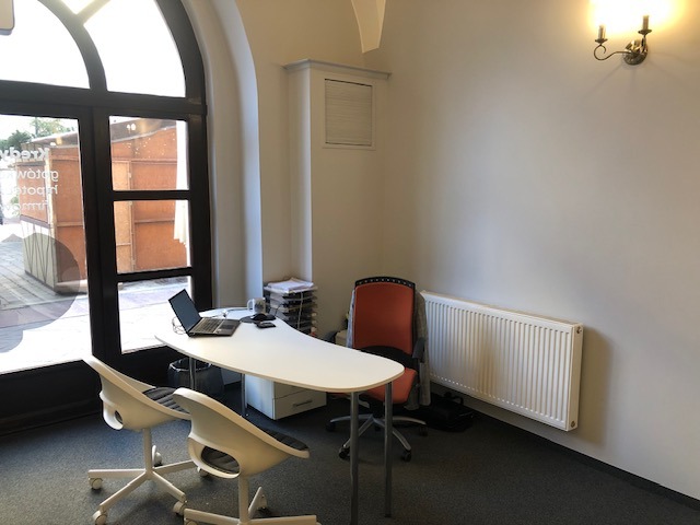 Jasne biuro z białym biurkiem, laptopem, dwoma nowoczesnymi krzesłami, pomarańczowym krzesłem biurowym, grzejnikiem i lampą ścienną w stylu retro.