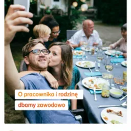 Mężczyzna w okularach robi selfie z kobietą, która go całuje w policzek na tle grupy osób siedzących przy stole z resztkami jedzenia i pustymi kieliszkami w ogrodzie. Logo Nationale Nederlanden...