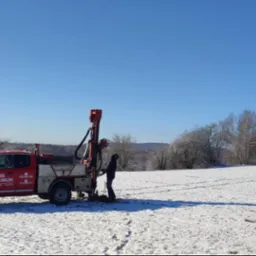 Czerwony samochód z wiertnicą na zasypanym śniegiem polu. Pracownik geologiczny w trakcie wiercenia. Błękitne niebo i zalesione wzgórza w tle.