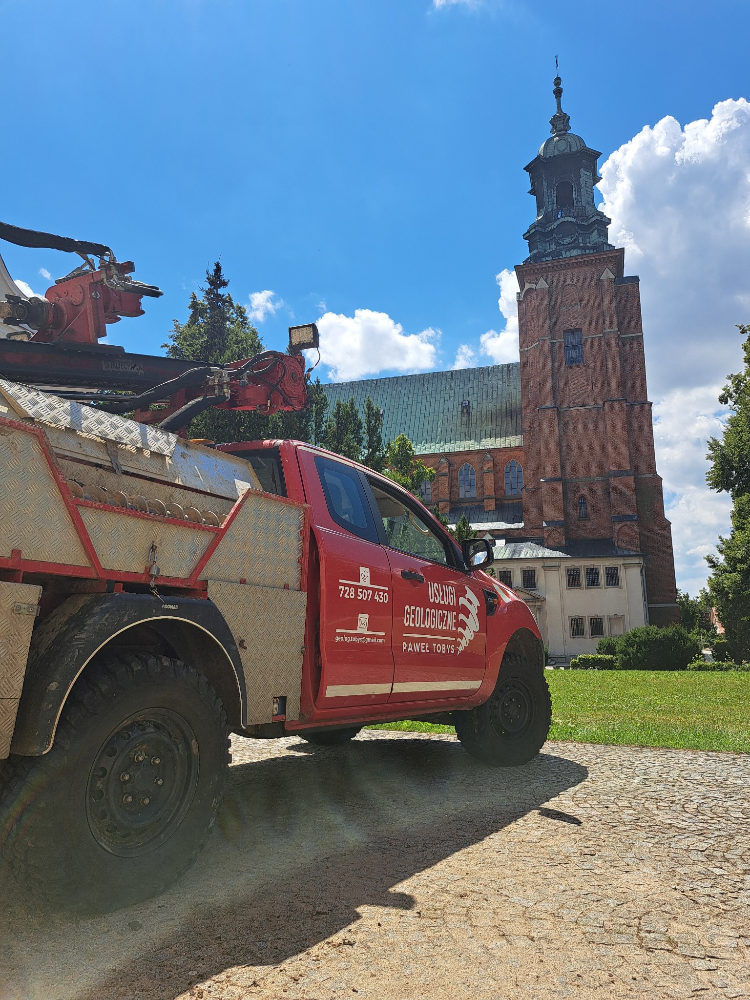 Czerwony samochód terenowy z wiertnicą firmy geologicznej na tle ceglanej wieży kościelnej w słoneczny dzień. Widoczne logo i numer telefonu firmy.