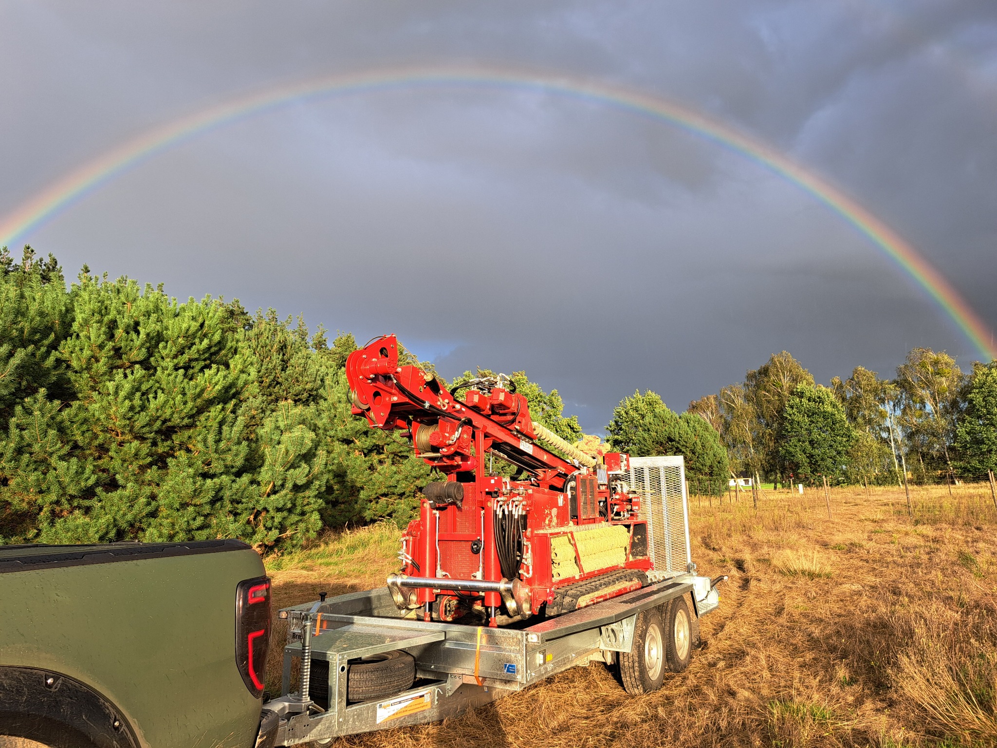 Czerwona wiertnica geologiczna na przyczepie, podwieszona do zielonego pickupa, na tle łąki i tęczy na pochmurnym niebie.