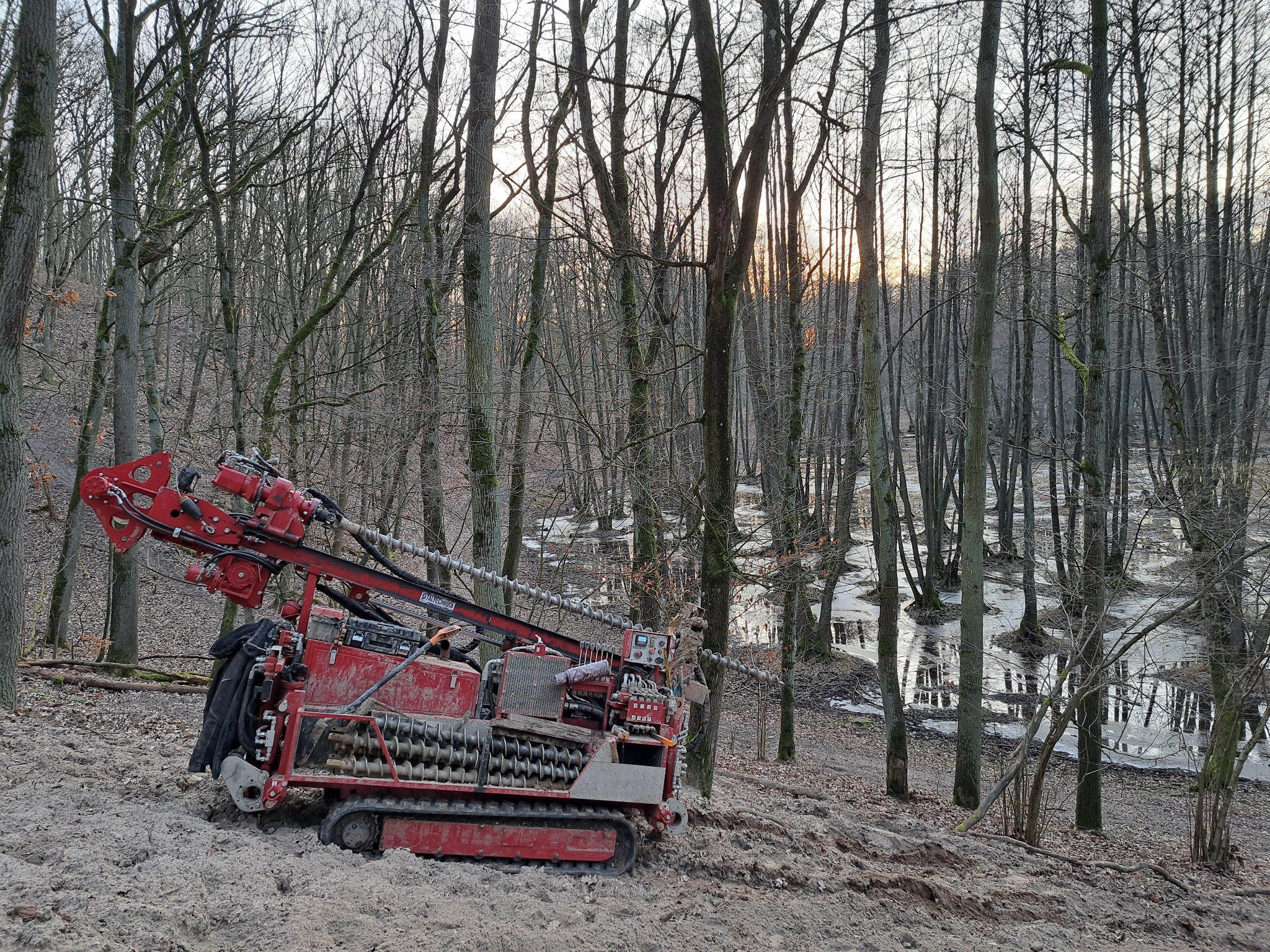 Czerwona wiertnica gąsienicowa w lesie, przygotowana do wiercenia, z widocznym świdrem. W tle drzewa i woda.