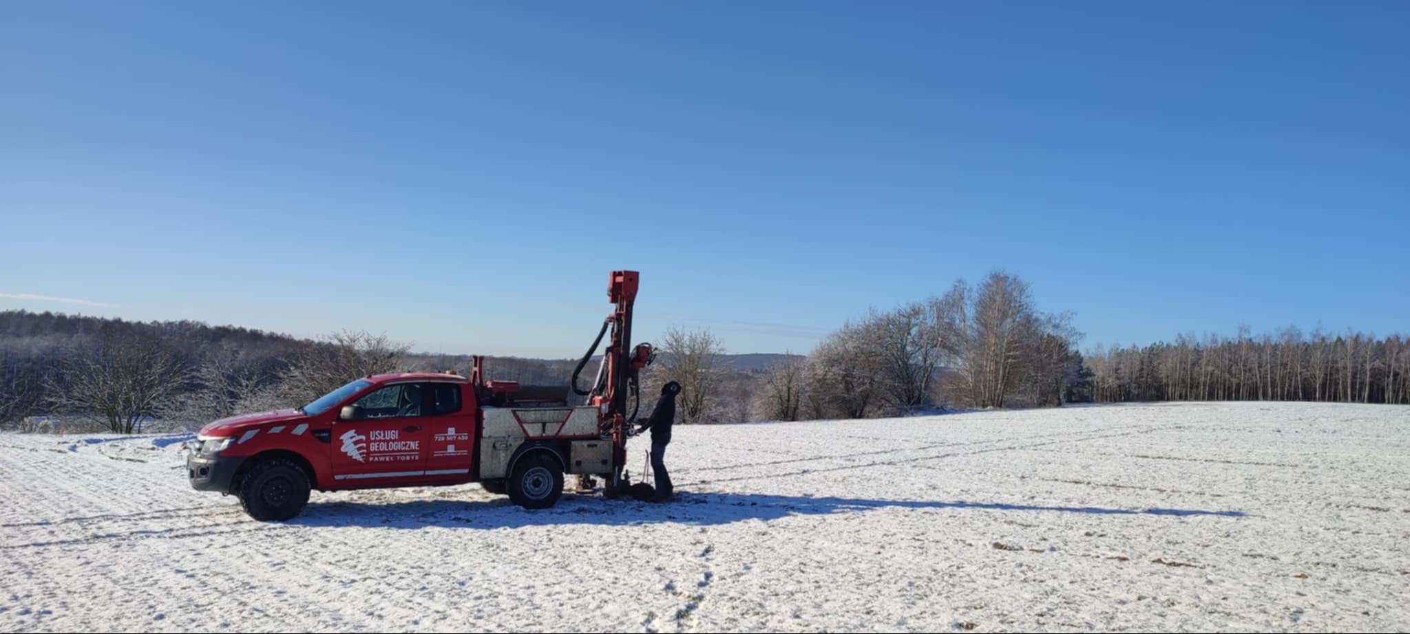Czerwony samochód z wiertnicą na zasypanym śniegiem polu. Pracownik geologiczny w trakcie wiercenia. Błękitne niebo i zalesione wzgórza w tle.