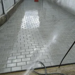 Świeżo ułożona, mokra szara kostka brukowa, spłukiwana wodą z węża ogrodowego, widoczne napięte żółte sznurki wyznaczające linię, betonowy płot w tle.