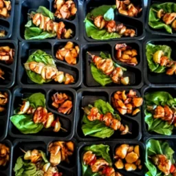 Gotowe zestawy cateringowe w czarnych pojemnikach z szaszłykami z kurczaka i pieczonymi ziemniakami, ułożone na liściach sałaty.