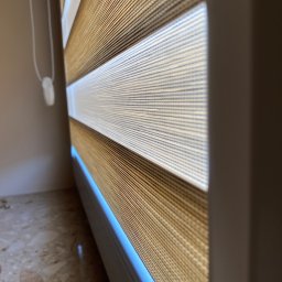 VERRANO Piotr Bandrowski - Zaciemniająca osłona okienna z naprzemiennymi pasami w kolorze beżowym i białym, widoczna z bliska, z mechanizmem łańcuszkowym po lewej stronie.