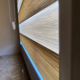 Zaciemniająca osłona okienna z naprzemiennymi pasami w kolorze beżowym i białym, widoczna z bliska, z mechanizmem łańcuszkowym po lewej stronie.