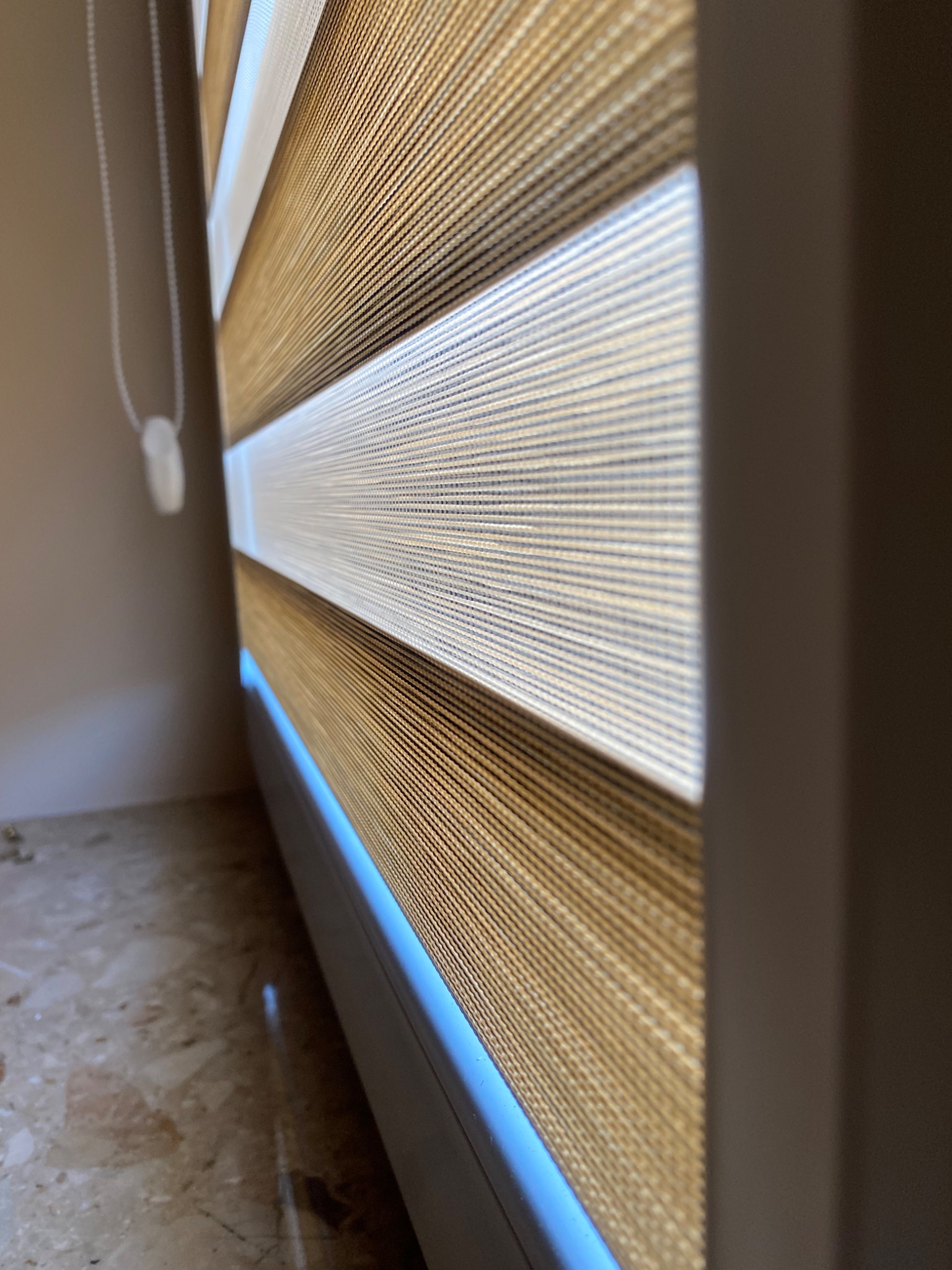 Zaciemniająca osłona okienna z naprzemiennymi pasami w kolorze beżowym i białym, widoczna z bliska, z mechanizmem łańcuszkowym po lewej stronie.