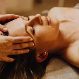 Kobieta z zamkniętymi oczami relaksuje się podczas masażu głowy wykonywanego w Rybienko Nowe. Uwagę przyciąga srebrny kolczyk w kształcie psa w uchu klientki.