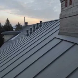 Szary dach z blachy na rąbek stojący z kominem z szarej cegły i kilkoma odpowietrznikami, widok z góry pod kątem.