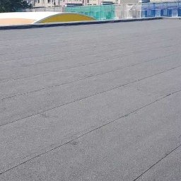 Bud-Henryk - Szary, płaski dach pokryty papą termozgrzewalną z widocznymi łączeniami, na tle częściowo widocznych budynków.